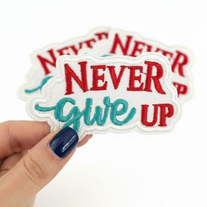 May include: An embroidered patch with a white background and a scalloped edge. The text NEVER is stitched in red, while the word Give is in teal script, and the word UP is in red. The patch features a layered design with multiple identical pieces.