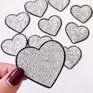 May include: A collection of heart-shaped patches featuring silver sequins on the front surface. Each heart is outlined with a solid black border. The patches are arranged in a group, with one heart held up in the foreground to show detail.