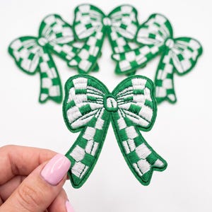 May include: A set of embroidered patches shaped like bows, featuring a green and white checkered pattern. The bows have a distinct knot in the center and two trailing ribbon ends. The embroidery is dense, creating a textured surface.