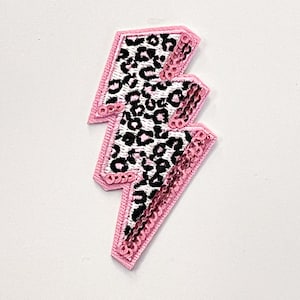 May include: Pink and white lightning bolt shaped patch with a leopard print design. The patch is made of sequins and is perfect for adding a touch of sparkle to any project.