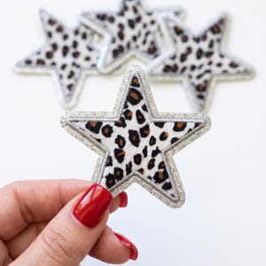 May include: A set of star-shaped patches featuring a white base with a black and brown leopard print pattern. Each star is outlined with a silver glitter border. The patches are arranged with one in the foreground and three in the background.