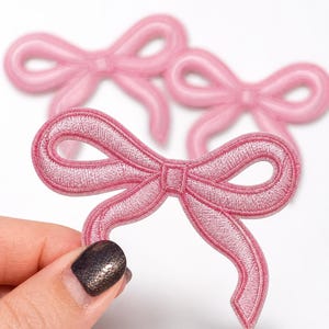 May include: Three pink embroidered bow patches featuring a satin-like thread finish. Each patch is shaped like a classic tied ribbon with two loops and two trailing ends. The embroidery is dense and uniform, creating a raised texture on the surface.
