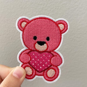 May include: Embroidered patch of a pink teddy bear holding a heart. The bear has a white outline, black eyes, and a tan snout and paws. The heart is pink with white polka dots. This patch is suitable for clothing or accessories.