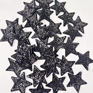 May include: A collection of black sequined star-shaped patches. Each star is densely covered in shiny black sequins, creating a textured, glittering surface. The patches are arranged in a scattered pattern, suitable for embellishing clothing or crafts.