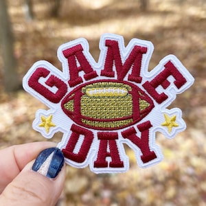 May include: Red and white embroidered patch with the words "GAME DAY" and a gold football in the center. The patch has two gold stars on either side of the football.