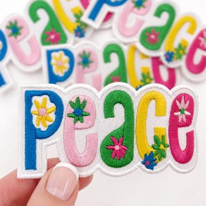 May include: A collection of embroidered fabric patches featuring the word Peace in various colors including blue, pink, green, and yellow. Each letter is decorated with small floral motifs. The patches have a white border and a textured finish.