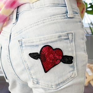 May include: Light blue denim jeans with a red sequined heart patch with a black arrow through it on the back pocket.