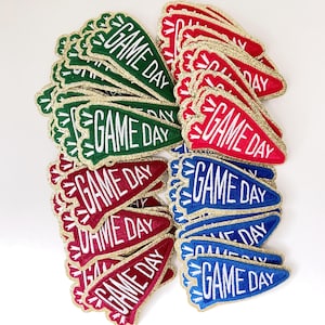 May include: A stack of felt pennant patches with gold glitter trim. The patches are red, green, and blue and each says "GAME DAY" in white lettering.