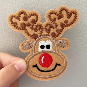 May include: A felt Christmas reindeer applique with brown antlers, a red nose, and a smiling mouth. The reindeer's eyes are black and white. The applique is held against a light gray background.