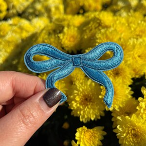 May include: A teal blue fabric bow with a satin finish. The bow is held in a hand against a background of yellow flowers.