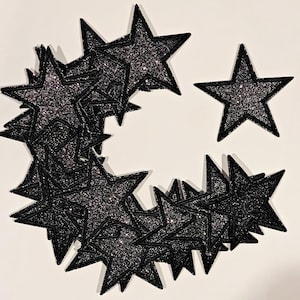 Puede incluir: Un gran grupo de parches en forma de estrella negra con purpurina, con un parche de estrella más pequeño a la derecha.