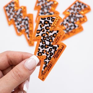 May include: A set of four lightning bolt-shaped patches featuring a white background with a black and orange leopard print pattern. Each patch is outlined with bright orange sequins that catch the light, creating a textured and reflective surface.