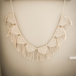 May include: A white macrame garland with triangle shapes and tassels hangs from a wooden rack with hooks.
