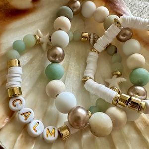 May include: Three layered bracelets with white, beige, and green beads. The bottom bracelet has the word "CALM" spelled out in gold letters on white beads.