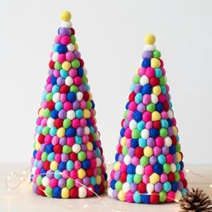May include: Two colorful, cone-shaped decorative trees. Each tree is covered in a variety of small, round, felt balls in shades of red, pink, blue, green, yellow, and purple. A small yellow ball tops each tree. The trees are on a light-colored surface.