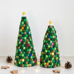 May include: Two decorative Christmas trees made of green, brown, and colorful pom-poms. The trees are cone-shaped with a yellow ball on top. Small wooden stars, pine cones, and string lights are scattered around the base of the trees.