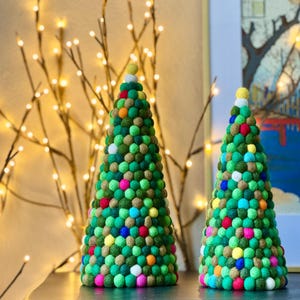 May include: Two decorative Christmas trees made of green felt balls in varying shades, adorned with colorful accents. The trees are cone-shaped and sit on a dark surface, with a backdrop of fairy lights and a framed picture.