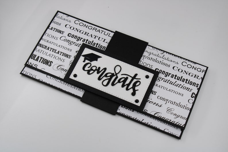 Pu&ograve; includere: Biglietto di laurea bianco e nero con un cappello di laurea nero e la parola "congrats" in corsivo nero. Il biglietto &egrave; piegato e ha un nastro nero.