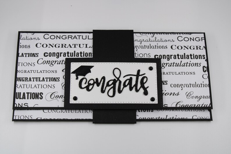 Pu&ograve; includere: Un biglietto di laurea bianco e nero con un cappello di laurea nero e la parola "congrate" in corsivo nero. Il biglietto &egrave; decorato con un motivo bianco e nero della parola "congratulazioni".