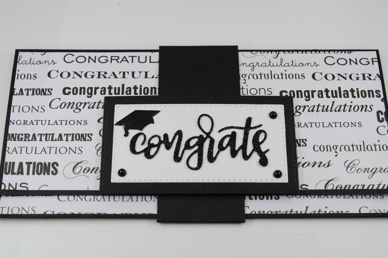 Pu&ograve; includere: Biglietto di laurea bianco e nero con un cappello di laurea nero e la parola "congrats" in lettere glitter nere.