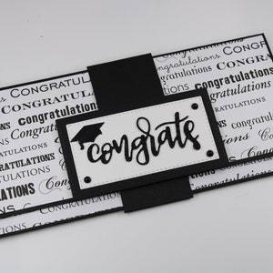 May include: Black and white graduation card with a black graduation cap and the word "congratulate" written in black glitter. The card is folded in half and has a black ribbon.