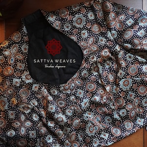 May include: A black and white printed blouse with a round neckline. The print features a repeating pattern of small circles, stars, and flowers. The blouse has a short sleeve and is made of a soft, lightweight fabric. The text "SATTVA WEAVES Timeless elegance" is printed on the front of the blouse.