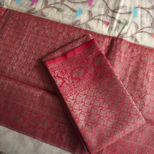 May include: A folded red textile with intricate patterns and a contrasting red border. The fabric has a shimmering quality, suggesting a luxurious material. The background features a floral patterned fabric.