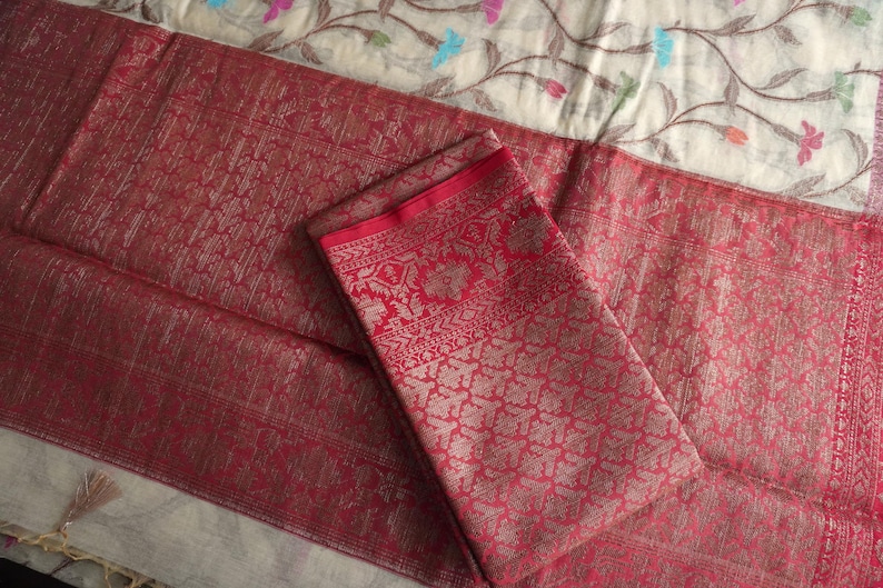 May include: A folded red textile with intricate patterns and a contrasting red border. The fabric has a shimmering quality, suggesting a luxurious material. The background features a floral patterned fabric.