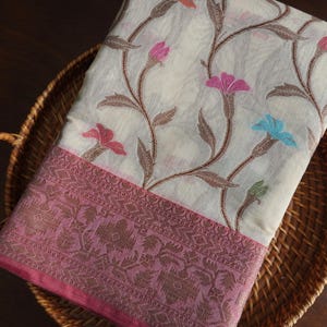 May include: A cream-colored textile with a floral pattern featuring pink, blue, and orange flowers and brown vines. The border is a darker pink with an intricate design. The textile is folded and placed in a woven basket.