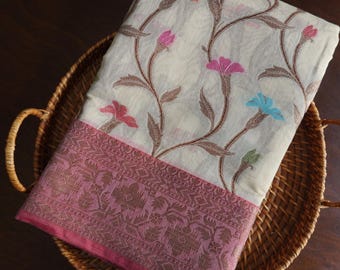 May include: A cream-colored textile with a floral pattern featuring pink, blue, and orange flowers and brown vines. The border is a darker pink with an intricate design. The textile is folded and placed in a woven basket.