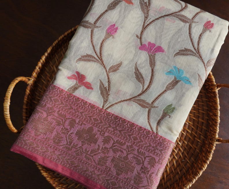May include: A cream-colored textile with a floral pattern featuring pink, blue, and orange flowers and brown vines. The border is a darker pink with an intricate design. The textile is folded and placed in a woven basket.
