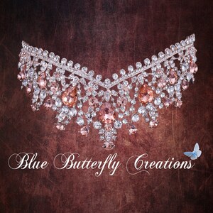 Pode incluir: Um colar delicado de prata e cristal com um design de lágrima e strass rosa. O colar está sobre um fundo marrom texturizado com o texto "Blue Butterfly Creations" e uma borboleta azul.