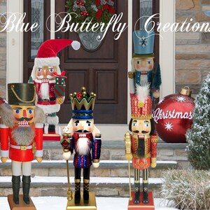 May include: Four wooden nutcracker figurines stand on a porch decorated for Christmas. The nutcrackers are dressed in red, blue and gold costumes. The nutcrackers are holding various props, including a sword, a sceptre and a drum. The nutcrackers are standing in front of a door with a wreath and a sign that reads "Blue Butterfly Creations".