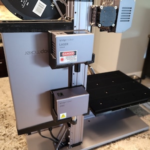 Puede incluir: Una impresora 3D Snapmaker gris con un cabezal láser y un cabezal CNC. El cabezal láser tiene una etiqueta de advertencia que dice "DANGER 1600mW".