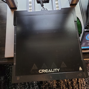 May include: A black 3D printer bed with a warning sign that says "Do Not Touch the Surface" and the CREALITY logo.