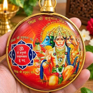 May include: A round gold-toned metal pendant featuring a colorful image of the five-faced deity Hanuman. The design includes a blue and red geometric yantra pattern with Sanskrit text and numerals, topped with a circular gold hanging loop.