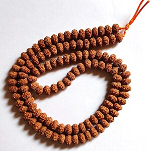 Può includere: Una collana di perle mala marrone con un cordoncino arancione annodato. Le perle sono fatte di materiali naturali e hanno una texture ruvida.