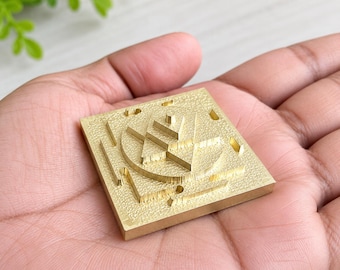 Puede incluir: Una placa cuadrada de latón con una superficie texturizada y un diseño geométrico en relieve. El centro muestra un motivo circular que contiene formas triangulares y un pequeño pilar central, todo en un tono dorado metálico.