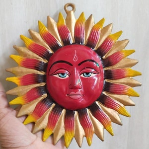 May include: A red sun with a human face painted on it. The sun has a gold, red, and black halo with yellow accents.