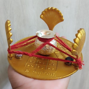 Könnte beinhalten: Ein goldfarbener Metallständer mit einem silbernen Topf in der Mitte. Der Ständer hat eine runde Basis mit einem Gittermuster und drei goldfarbene Metallblätter. Der Topf ist mit einer roten Schnur gebunden. Der Ständer ist mit dem Text "श्री" in Hindi verziert.