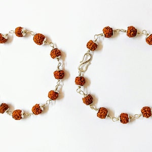 Peut inclure: Deux bracelets composés de perles rudraksha sphériques, brunes et texturées, reliées par un fil métallique argenté. Chaque bracelet comprend un fermoir à crochet en métal argenté et de petites entretoises argentées entre les perles.