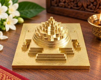 May include: A gold-colored metal Sri Yantra sculpture featuring a square base with a central three-dimensional stepped pyramid design composed of multiple concentric triangles and geometric layers, creating a complex, symmetrical, and tiered structure.