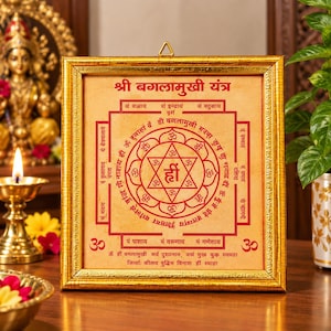 Könnte beinhalten: Ein quadratischer Wandrahmen mit Goldfinish, der einen beigen Druck des Shri Baglamukhi Yantra enthält. Das Design zeigt rote geometrische Muster, Sanskrit-Text und das Om-Symbol in einem Stern, alles durch Glas geschützt.