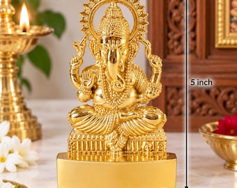 May include: A gold-colored Ganesha figurine in a seated position, featuring intricate detailing on the crown, jewelry, and clothing. The statue is mounted on a rectangular base and includes a vertical measurement line indicating a height of 5 inches.