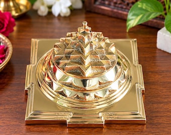May include: A polished gold-colored metal Sri Yantra pyramid sculpture featuring a multi-layered geometric design with intricate triangular patterns rising to a central point, set upon a square base with stepped edges and a reflective metallic finish.