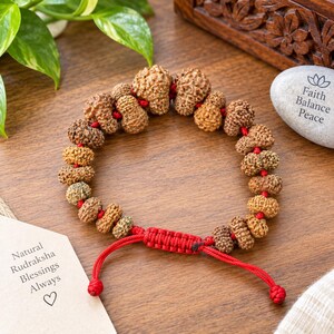 May include: A bracelet made of natural brown, textured Rudraksha beads strung on a bright red cord. The bracelet features an adjustable sliding knot closure with two dangling red cord ends. The beads are round with a rough, organic surface.