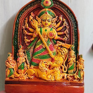 Puede incluir: Una escultura en relieve de terracota pintada a mano que representa a una diosa hindú con múltiples brazos, de pie sobre un león. La diosa está adornada con intrincadas joyas y una corona. La escultura está enmarcada en un arco marrón con acentos verdes y dorados.