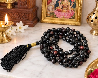 May include: A prayer bead necklace featuring smooth, dark brown and black oval-shaped beads strung together. The strand is finished with a long, black thread tassel secured by a metallic gold-colored thread wrapping at the top of the tassel.