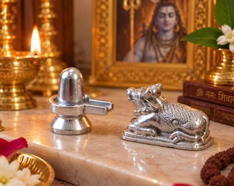 Könnte beinhalten: Ein silberfarbener Shiva Lingam und eine silberfarbene Nandi-Stier-Figur. Der Shiva Lingam hat eine glatte, abgerundete Oberseite und einen zylindrischen Sockel. Der Nandi-Stier ist in liegender Position mit detaillierten Gravuren dargestellt.