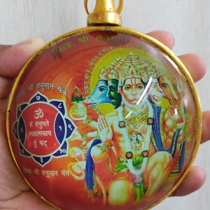 May include: A round, gold-framed, glass ornament with a red background and a colorful image of the Hindu deity Hanuman. The image includes Sanskrit text and the words "Shree Hanuman Yantra".